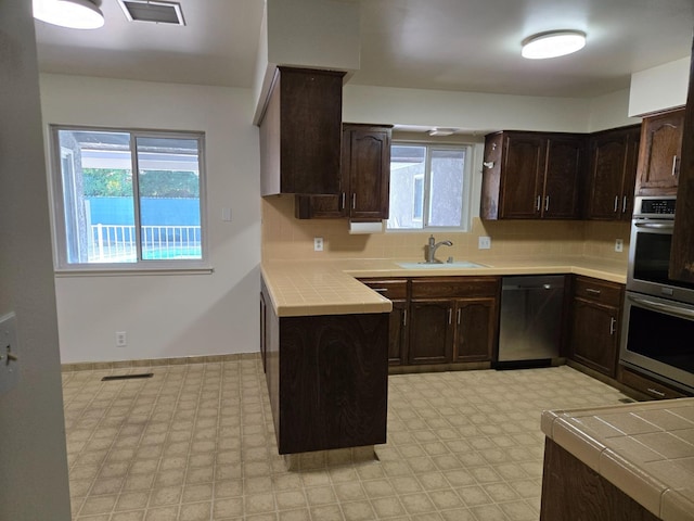 kitchen featuring a sink, appliances with stainless steel finishes, light floors, dark brown cabinets, and tile counters