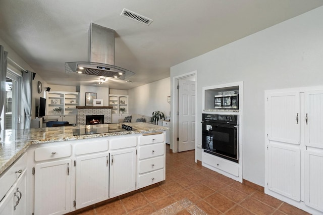 kitchen featuring visible vents, black appliances, white cabinets, island range hood, and a brick fireplace