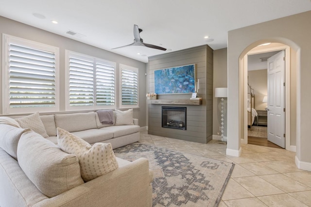 living area featuring a ceiling fan, visible vents, light tile patterned flooring, arched walkways, and a glass covered fireplace