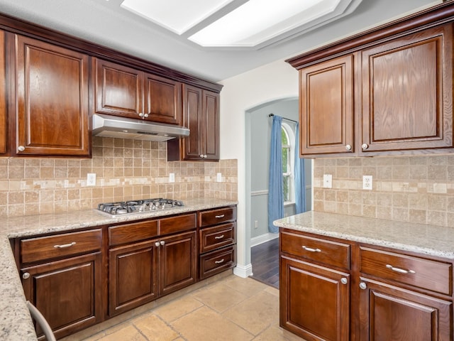 kitchen with backsplash, light stone countertops, under cabinet range hood, stainless steel gas stovetop, and arched walkways