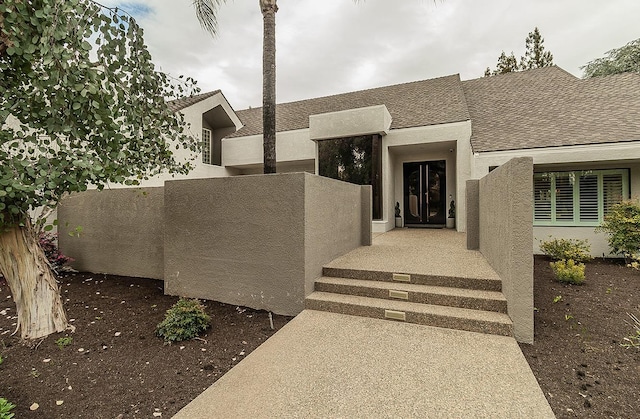 view of exterior entry with a shingled roof, fence, and stucco siding