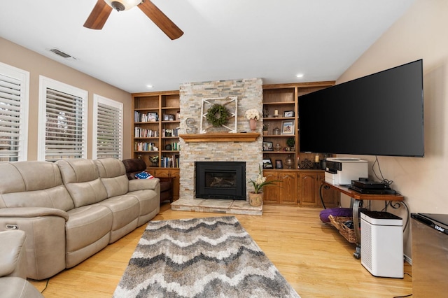 living room with built in shelves, a ceiling fan, visible vents, a stone fireplace, and light wood-style floors