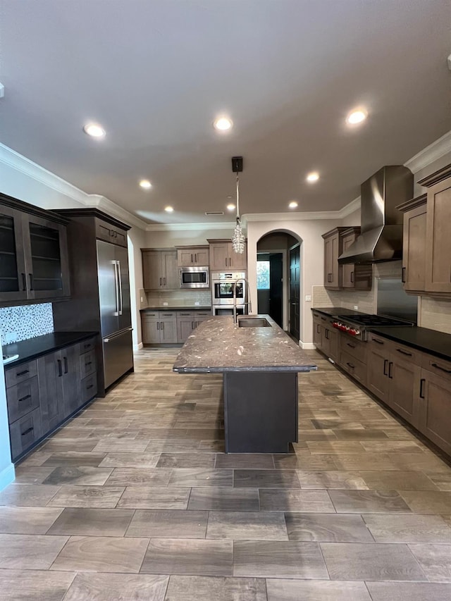 kitchen with decorative backsplash, built in appliances, arched walkways, and wall chimney exhaust hood