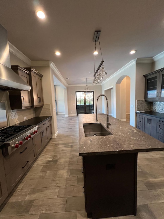 kitchen with arched walkways, a kitchen island with sink, a sink, stainless steel gas stovetop, and wall chimney range hood