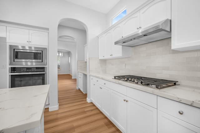 kitchen featuring light wood-type flooring, under cabinet range hood, white cabinetry, arched walkways, and appliances with stainless steel finishes