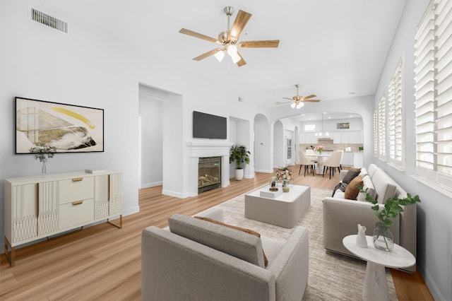 living room featuring visible vents, light wood-type flooring, arched walkways, a glass covered fireplace, and a ceiling fan