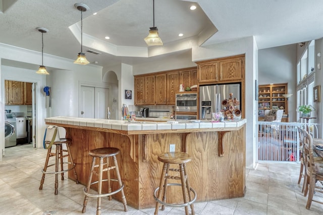 kitchen with tile countertops, washer / clothes dryer, brown cabinetry, and appliances with stainless steel finishes