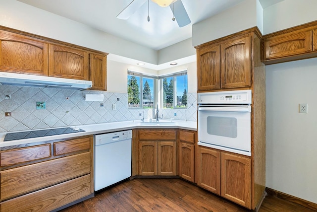 kitchen with under cabinet range hood, a sink, backsplash, white appliances, and dark wood-style flooring