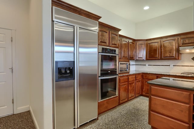 kitchen featuring backsplash, stainless steel appliances, exhaust hood, and dark stone countertops