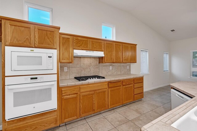 kitchen featuring white appliances, tile countertops, light tile patterned flooring, decorative backsplash, and under cabinet range hood