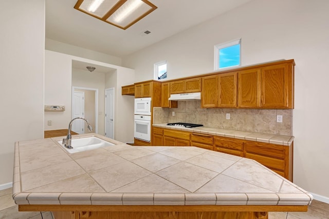 kitchen featuring white appliances, a sink, decorative backsplash, under cabinet range hood, and brown cabinets