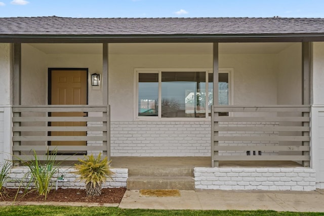 view of exterior entry featuring stucco siding, brick siding, covered porch, and a shingled roof