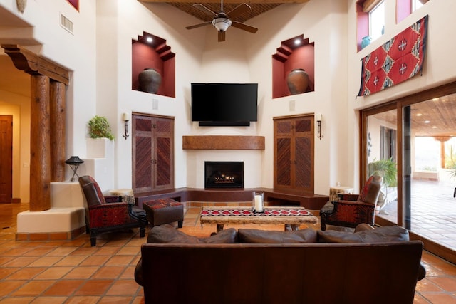 tiled living room with visible vents, a warm lit fireplace, a towering ceiling, decorative columns, and ceiling fan