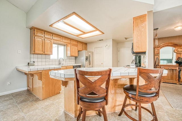 kitchen with a peninsula, a sink, tile counters, stainless steel refrigerator with ice dispenser, and tasteful backsplash