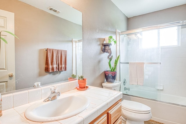 bathroom with vanity, visible vents, toilet, a textured wall, and combined bath / shower with glass door