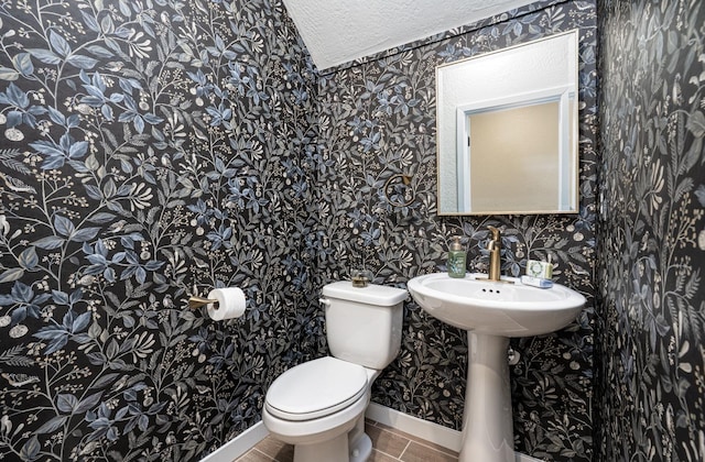 half bathroom featuring tile patterned floors, toilet, a textured ceiling, wallpapered walls, and baseboards