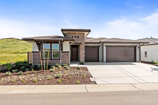 prairie-style home featuring a tile roof, driveway, a garage, brick siding, and stucco siding
