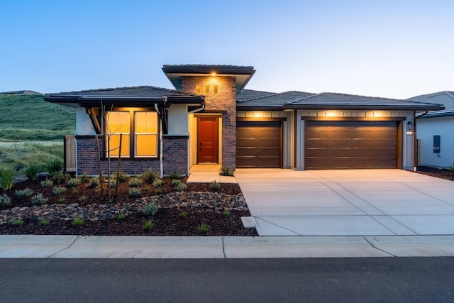 prairie-style home with a tiled roof, stucco siding, an attached garage, driveway, and brick siding