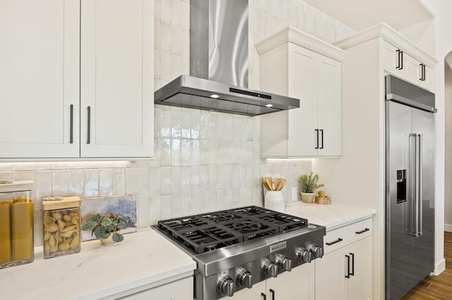 kitchen featuring light stone counters, backsplash, stainless steel appliances, white cabinets, and wall chimney range hood