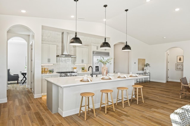 kitchen with wall chimney range hood, built in refrigerator, range, arched walkways, and a kitchen island with sink