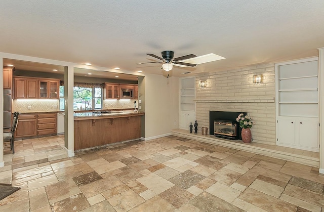 kitchen with a ceiling fan, brown cabinetry, open floor plan, stone tile flooring, and a brick fireplace