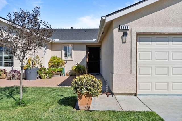 view of exterior entry featuring a yard, an attached garage, a shingled roof, and stucco siding