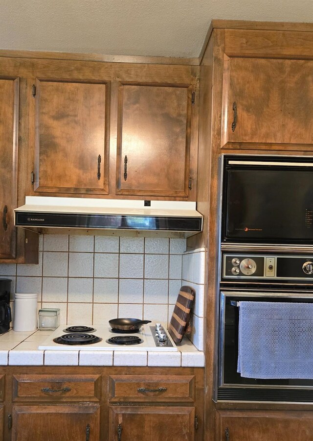 26174 Haley Way, Madera, CA 93638 - kitchen with brown cabinetry, under cabinet range hood, white electric stovetop, oven, and backsplash