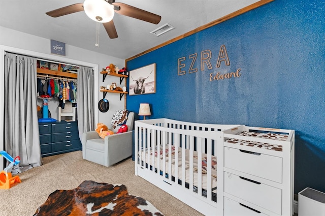 carpeted bedroom featuring a closet, visible vents, a crib, ceiling fan, and a textured wall