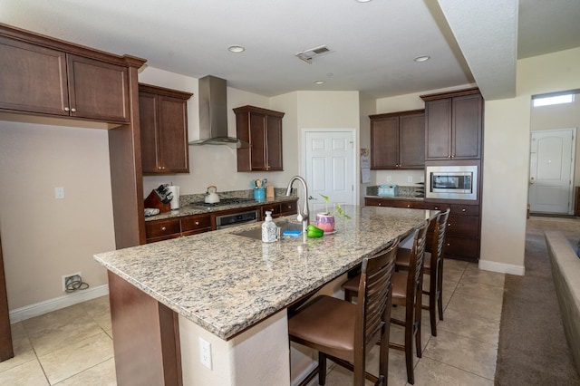 kitchen with wall chimney range hood, a sink, visible vents, stainless steel appliances, and a center island with sink