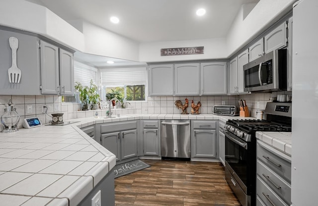 kitchen featuring stainless steel appliances, tile countertops, gray cabinetry, a sink, and dark wood-type flooring