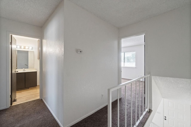 hallway with a textured ceiling, light colored carpet, a sink, an upstairs landing, and baseboards
