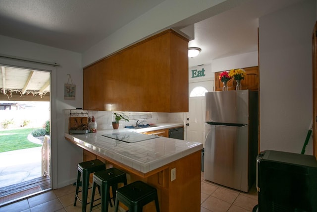 kitchen featuring freestanding refrigerator, a peninsula, light tile patterned flooring, tile counters, and brown cabinets