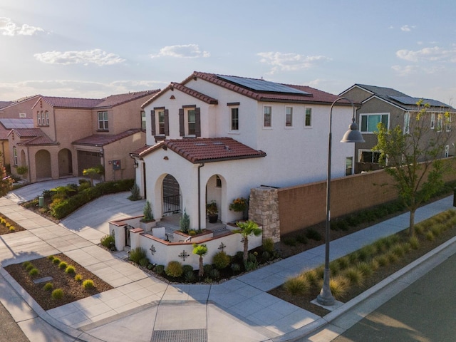mediterranean / spanish-style house with roof mounted solar panels, a tile roof, fence, concrete driveway, and a residential view