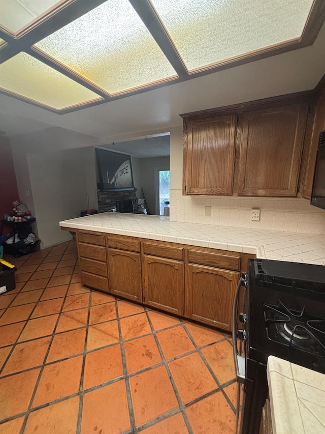 1225 Pontiac Ave, Clovis, CA 93612 - kitchen with black gas stove, tile countertops, brown cabinetry, a peninsula, and backsplash