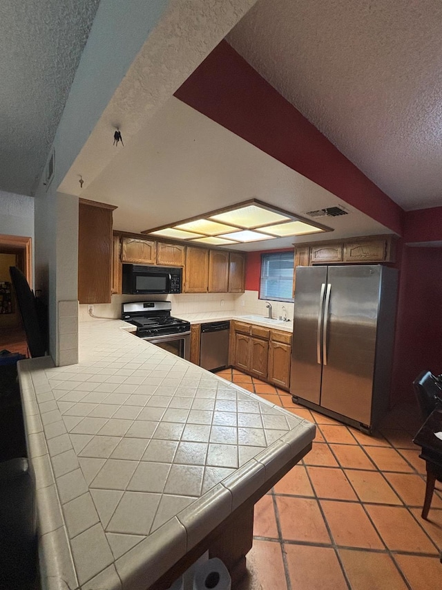1225 Pontiac Ave, Clovis, CA 93612 - kitchen with stainless steel appliances, a textured ceiling, tile countertops, a peninsula, and brown cabinetry