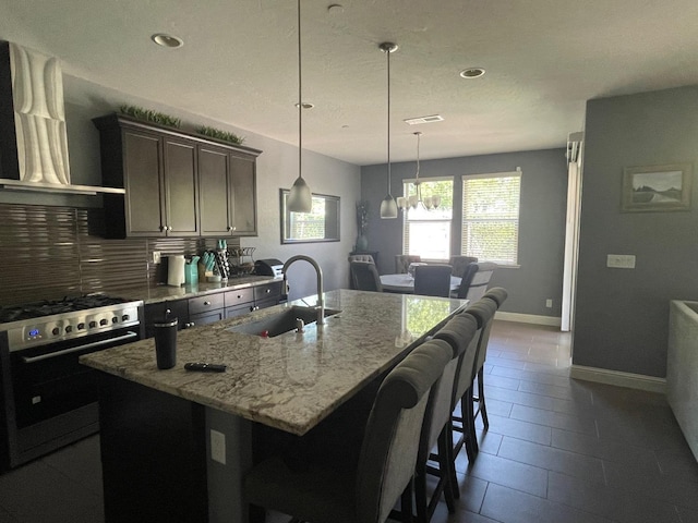kitchen featuring wall chimney range hood, light stone countertops, a kitchen island with sink, stainless steel range with gas stovetop, and a sink