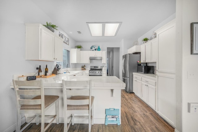 kitchen featuring tile counters, under cabinet range hood, stainless steel appliances, a sink, and a peninsula