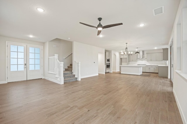 unfurnished living room featuring light wood finished floors, baseboards, visible vents, ceiling fan with notable chandelier, and stairs