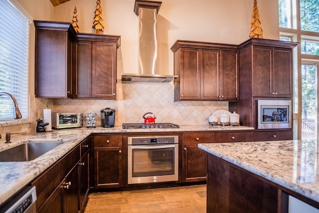 kitchen featuring light stone counters, a sink, decorative backsplash, stainless steel appliances, and wall chimney exhaust hood