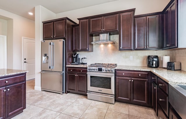kitchen with wall chimney exhaust hood, decorative backsplash, appliances with stainless steel finishes, light stone counters, and light tile patterned floors