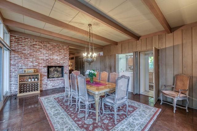 dining space featuring wood walls, an inviting chandelier, beam ceiling, dark tile patterned flooring, and a brick fireplace