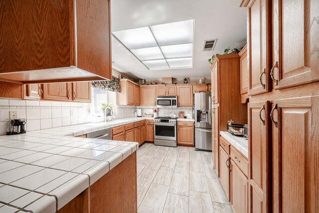 kitchen featuring tile counters, stainless steel appliances, light brown cabinets, a sink, and tasteful backsplash