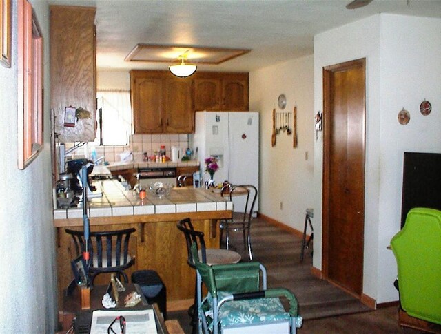 7081 Live Oak, Wonder Valley, CA 93657 - kitchen with tile counters, brown cabinets, dark wood-style floors, a peninsula, and white refrigerator with ice dispenser