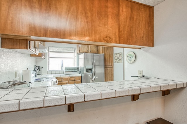 kitchen featuring tile counters, stainless steel fridge, brown cabinetry, and ventilation hood