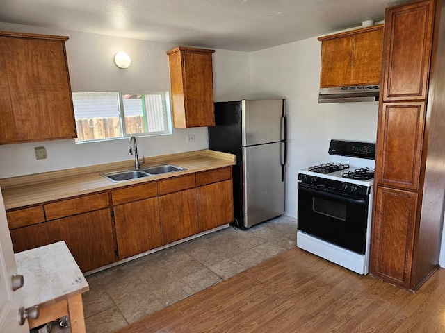 kitchen with a sink, freestanding refrigerator, under cabinet range hood, range with gas cooktop, and brown cabinetry