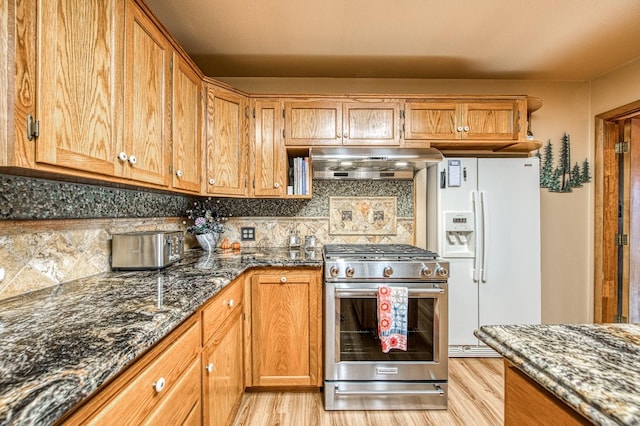 kitchen featuring dark stone countertops, white refrigerator with ice dispenser, under cabinet range hood, stainless steel gas range, and light wood finished floors