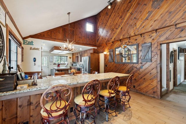 kitchen with light wood finished floors, an inviting chandelier, high vaulted ceiling, wooden walls, and stainless steel refrigerator with ice dispenser