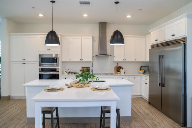 kitchen featuring visible vents, built in appliances, a kitchen island with sink, wall chimney range hood, and light countertops