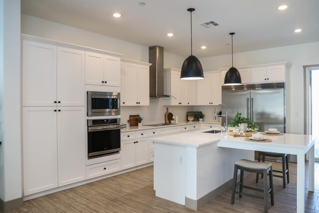 kitchen with visible vents, built in appliances, wall chimney range hood, white cabinets, and a sink