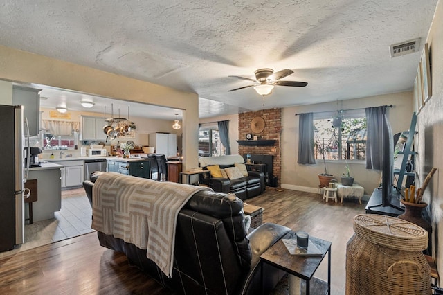 living room with ceiling fan, a textured ceiling, visible vents, a brick fireplace, and wood finished floors
